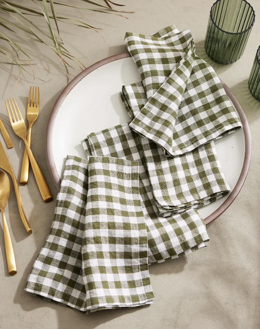 Four olive and white gingham napkins on a white ceramic plate next to some gold cutlery, green drinking glasses, and a plant