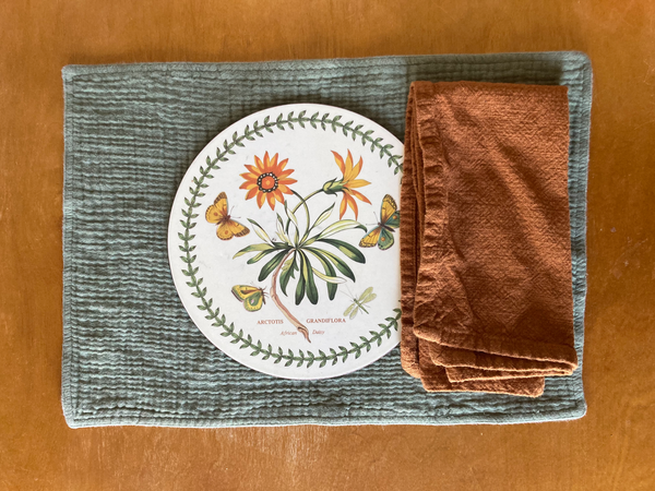 A table setting made of a green placemat, floral charger, and burnt orange cloth napkin sits on top of a wooden tabletop.