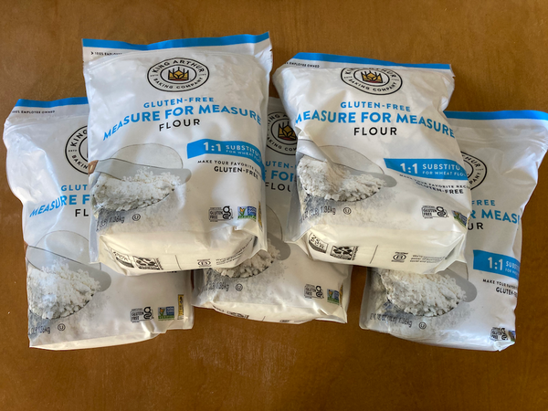 Five bags of King Arthur gluten-free measure for measure flour on a brown tabletop.