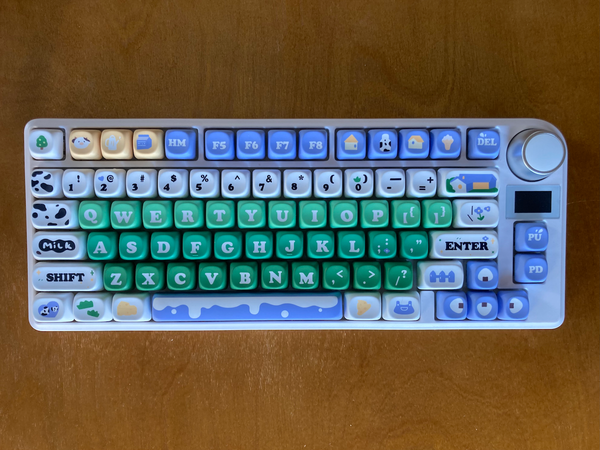 A keyboard with dairy farm-themed keycaps in green, white, blue, and yellow.